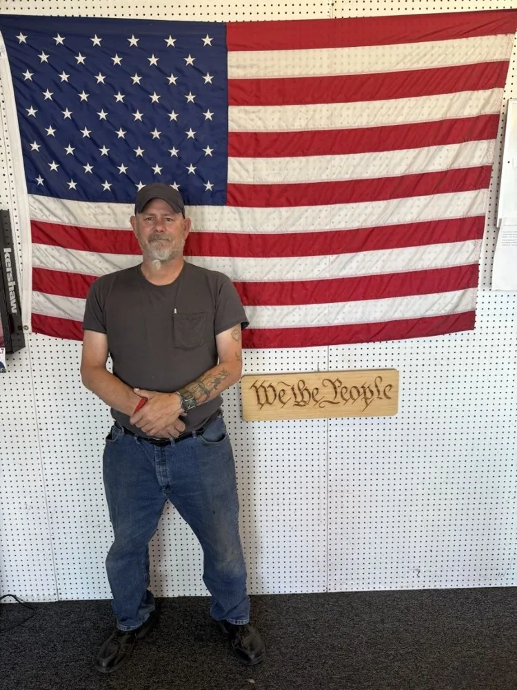 A team member standing in front of the shop's American flag and carved “We the People” sign
